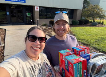 Two smiling individuals take a selfie outside Reynolds Consumer Products, holding boxes of food donations on a sunny day.