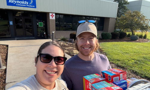 Two smiling individuals take a selfie outside Reynolds Consumer Products, holding boxes of food donations on a sunny day.