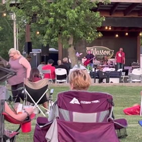Outdoor event with a stage performance; relaxed audience enjoying live music under cozy string lights in a green setting.