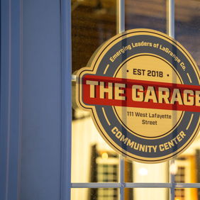 Sign for "The Garage" community center, established in 2018, framed by a window, evoking warmth and togetherness.