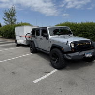 Parked gray Jeep Wrangler in a lot with a white trailer, greenery, and blue sky, evoking adventure and freedom.