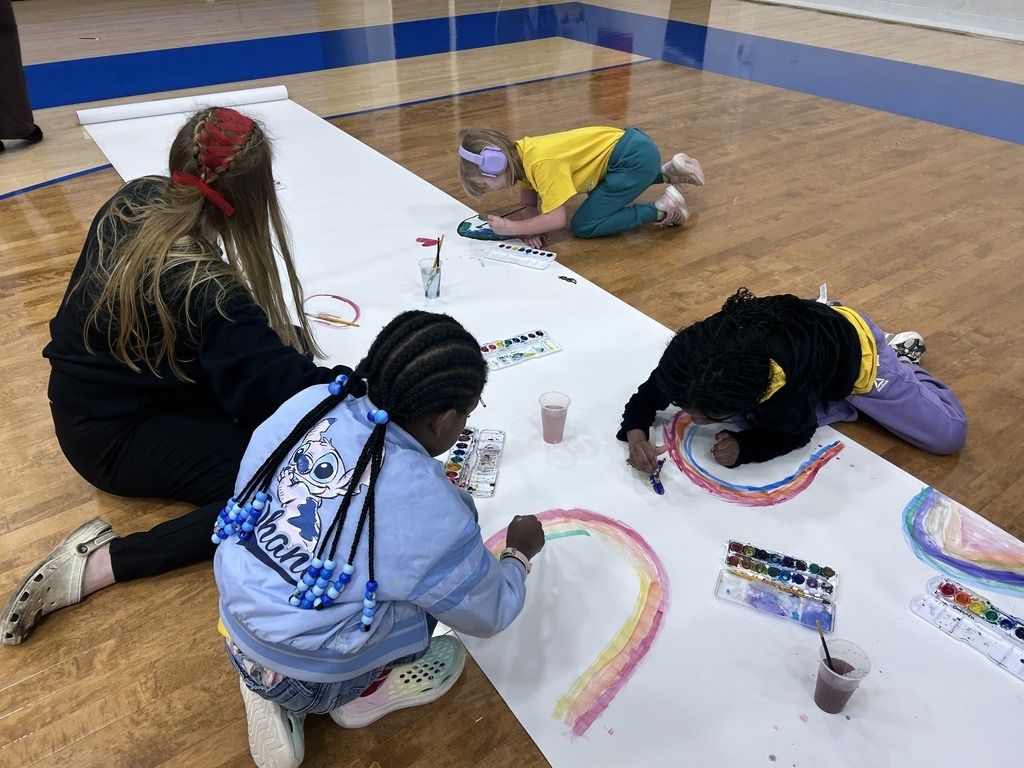 Children paint on a large paper roll, guided by an adult, in a joyful indoor setting filled with creativity and collaboration.
