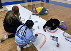 Children paint on a large paper roll, guided by an adult, in a joyful indoor setting filled with creativity and collaboration.
