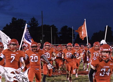 Orange-clad football players charge onto the field, flags in hand, embodying excitement and team spirit under a dusky sky.