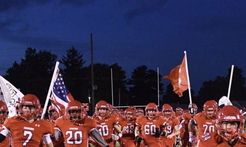 Orange-clad football players charge onto the field, flags in hand, embodying excitement and team spirit under a dusky sky.
