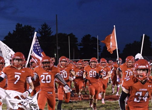 Orange-clad football players charge onto the field, flags in hand, embodying excitement and team spirit under a dusky sky.