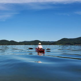 Serene outdoor scene with a calm lake, a distant kayaker, lush greenery, rolling hills, and a clear blue sky.