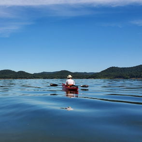 Serene outdoor scene with a calm lake, a distant kayaker, lush greenery, rolling hills, and a clear blue sky.