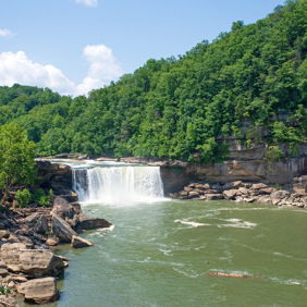 Waterfall cascading into a serene river, surrounded by greenery and hills under a bright blue sky, evokes tranquility and connection to nature.