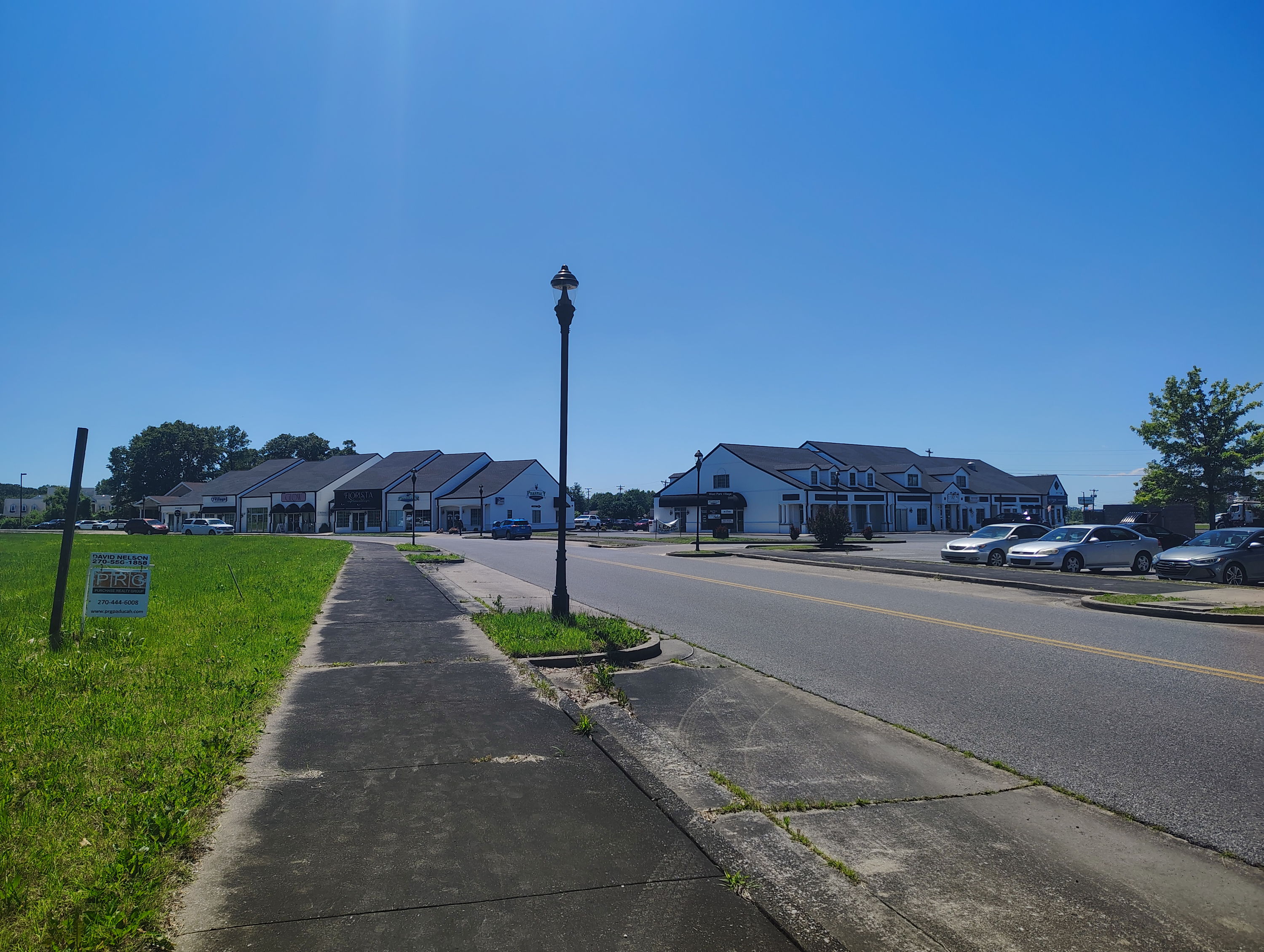 A sunny suburban scene with a clear sky, greenery, shops, and parked cars evokes calmness and tranquility.