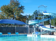 A blue swimming pool with a twisting water slide, surrounded by blue and white umbrellas and trees, evokes summer fun.