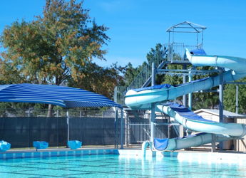 A blue swimming pool with a twisting water slide, surrounded by blue and white umbrellas and trees, evokes summer fun.