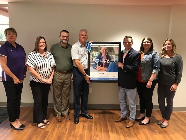 Group of eight joyfully celebrating an achievement, posing with a framed photo in a well-lit, professional setting.