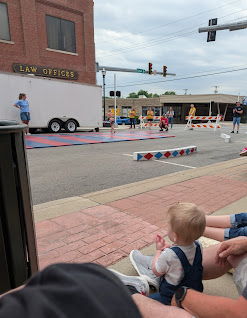 A child sits on the ground, enchanted by a lively community festival with activities and joyful people around them.