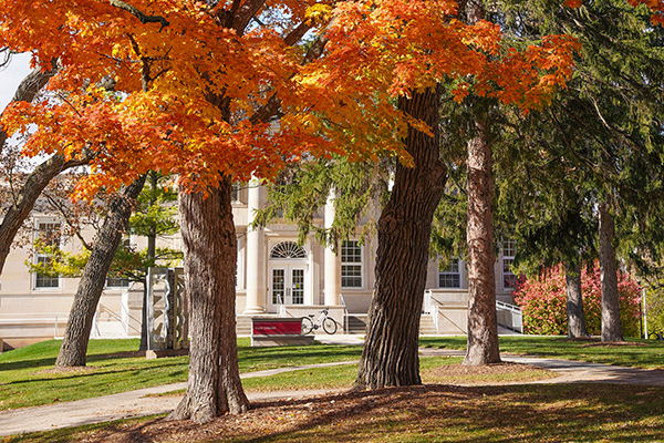 Autumn scene with vibrant orange and yellow leaves, a classic school building, lush green grass, and a winding path.