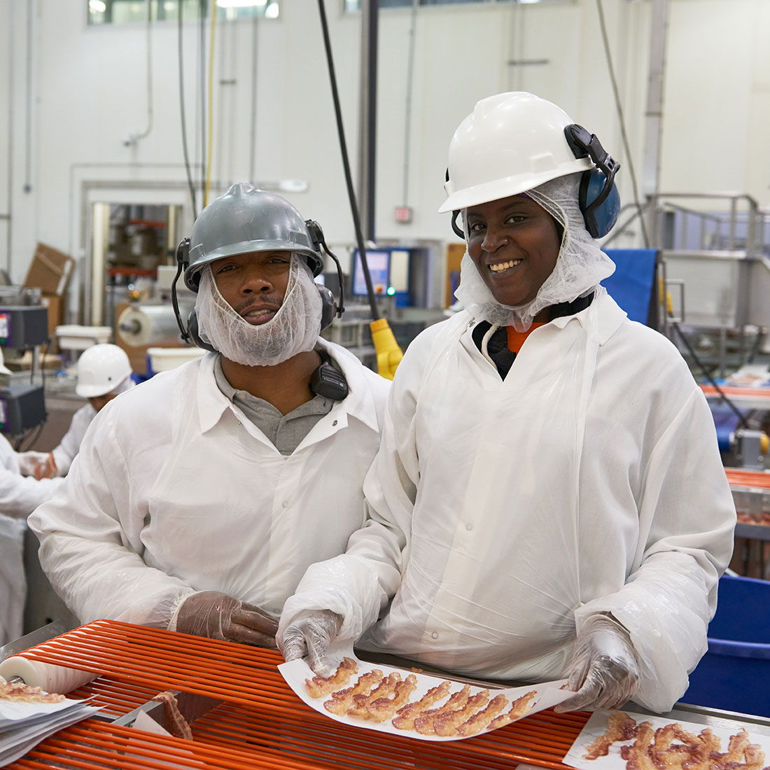 Two workers in protective gear smile while holding food trays in a professional, organized processing facility.