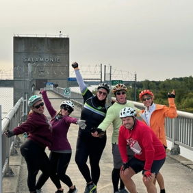 Six individuals in helmets and athletic wear smile on a bridge, exuding joy and camaraderie in a green recreational area.