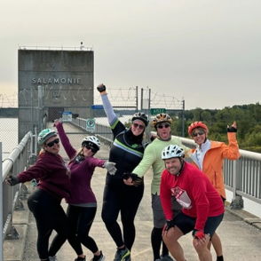 Six individuals in helmets and athletic wear smile on a bridge, exuding joy and camaraderie in a green recreational area.