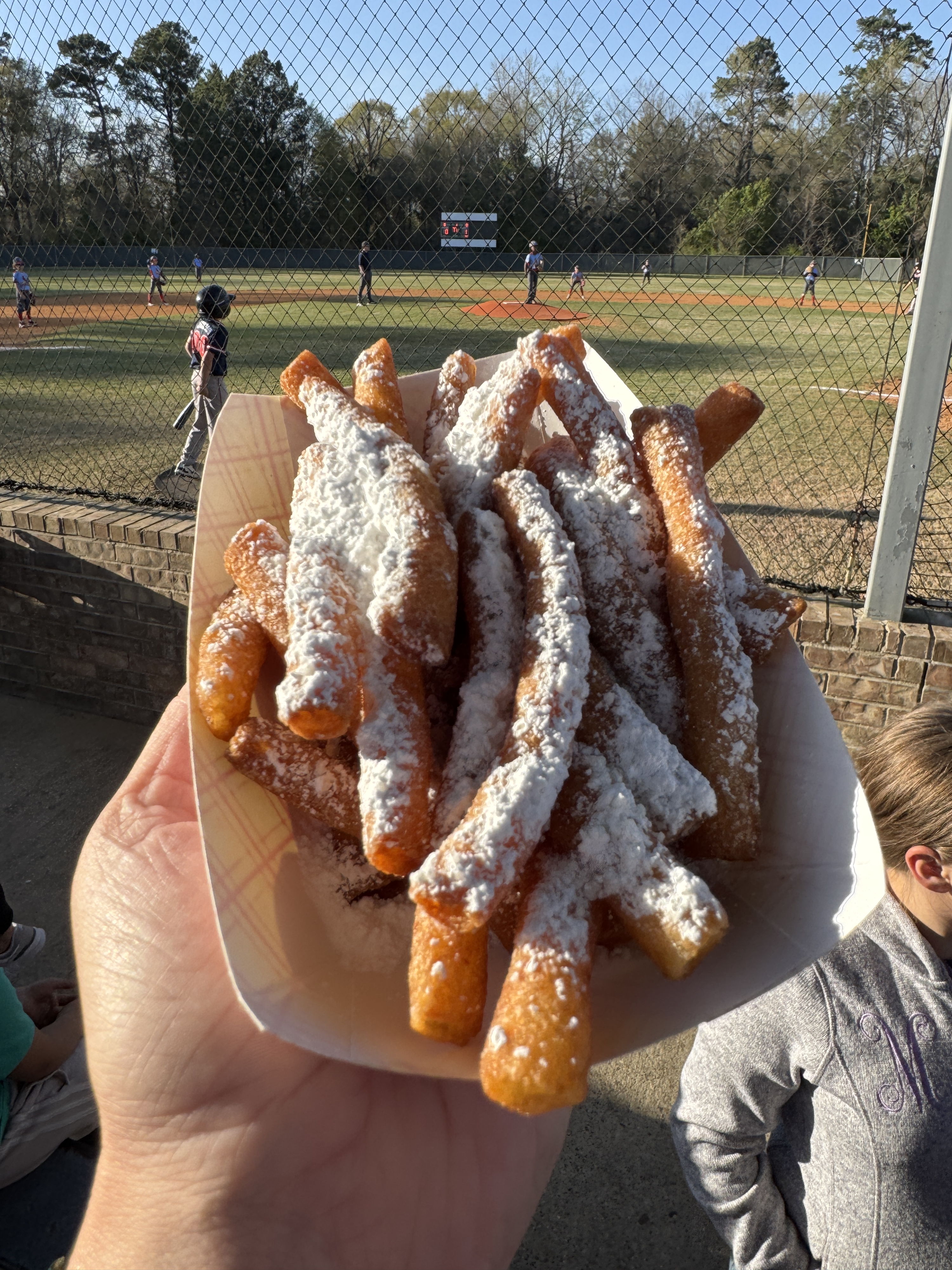 A hand holds funnel cake fries dusted with powdered sugar, set against a baseball field, evoking joy and nostalgia.