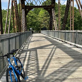 Rustic iron and wood bridge amidst greenery, with a blue bicycle inviting exploration under a bright sky and fluffy clouds.