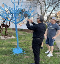 A blue tree with wooden hearts; a man adds a heart while another watches, symbolizing community and connection.