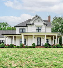Charming two-story house with white exterior, dark roof, and inviting porch, nestled in lush greenery under a cloudy sky.