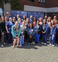 A large group of smiling people gathered outdoors, showcasing joy, unity, and camaraderie near a brick building.