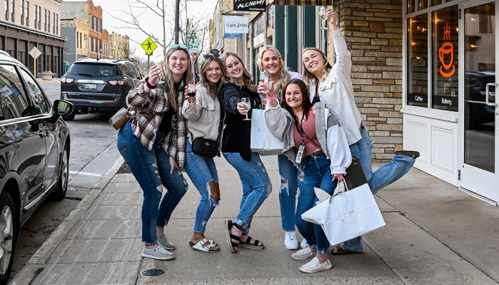 Six young women joyfully pose on a sidewalk in casual outfits, holding shopping bags, capturing a cheerful outing together.