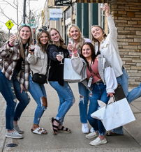 Six young women joyfully pose on a sidewalk in casual outfits, holding shopping bags, capturing a cheerful outing together.