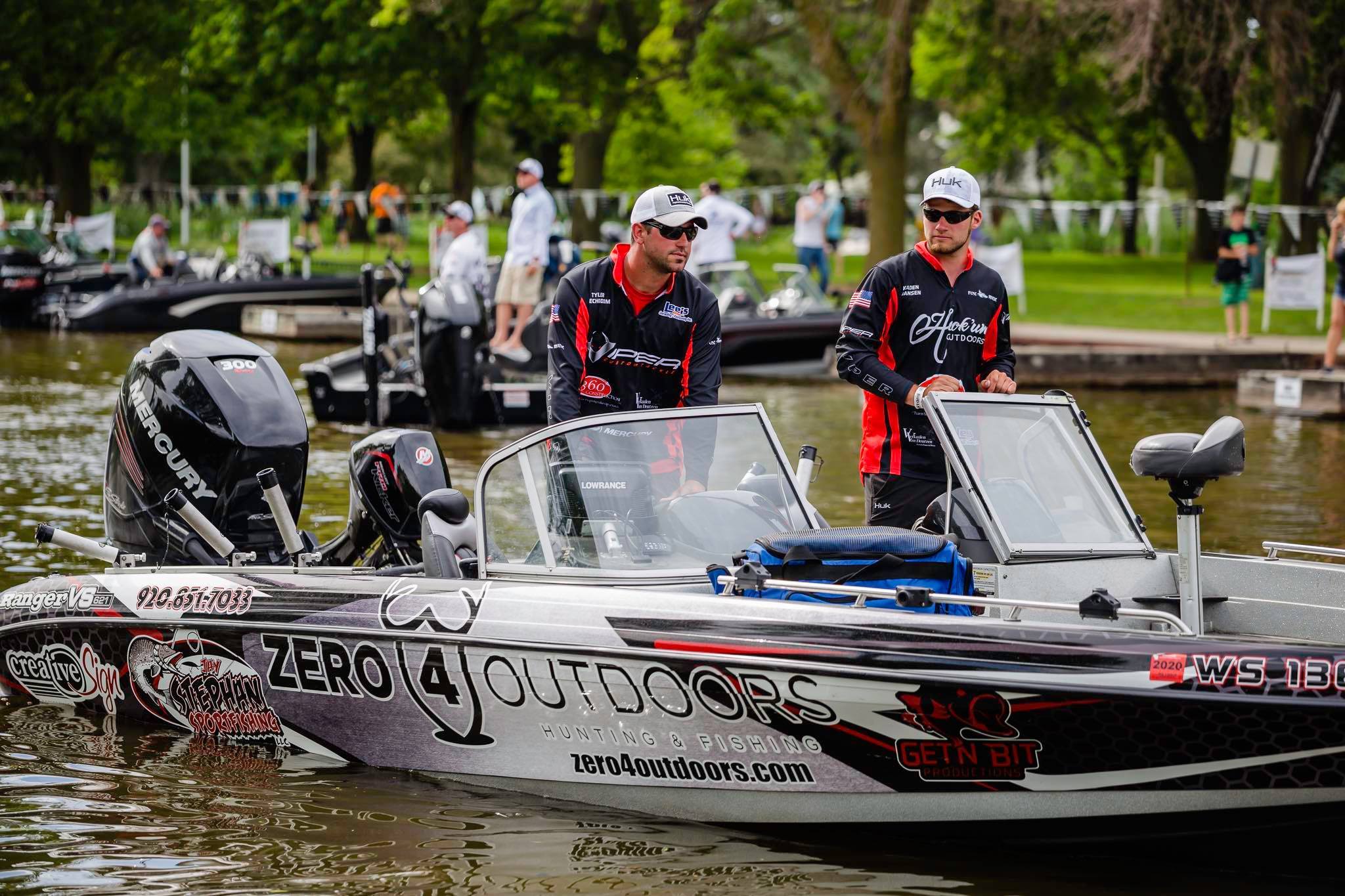 Two men in matching outfits on a well-equipped fishing boat, ready for a tournament in a serene, green outdoor setting.