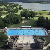 Bright, sunny pool day with people enjoying a vibrant blue pool surrounded by greenery and castle-like structures.