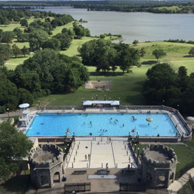 Bright, sunny pool day with people enjoying a vibrant blue pool surrounded by greenery and castle-like structures.