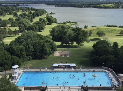 Bright, sunny pool day with people enjoying a vibrant blue pool surrounded by greenery and castle-like structures.
