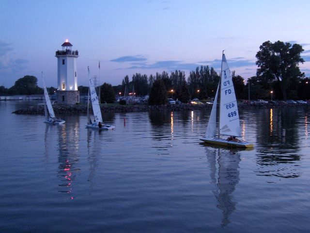 A serene evening scene with a lighthouse, sailboats, and a calm water reflection under a twilight sky of blues and purples.