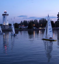 A serene evening scene with a lighthouse, sailboats, and a calm water reflection under a twilight sky of blues and purples.