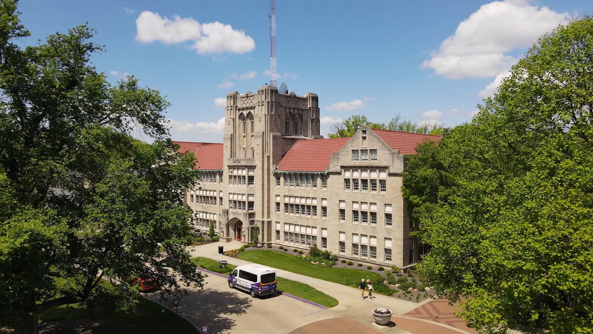 Large academic building with a stone facade and red roof, surrounded by green trees under a bright, partly cloudy sky.