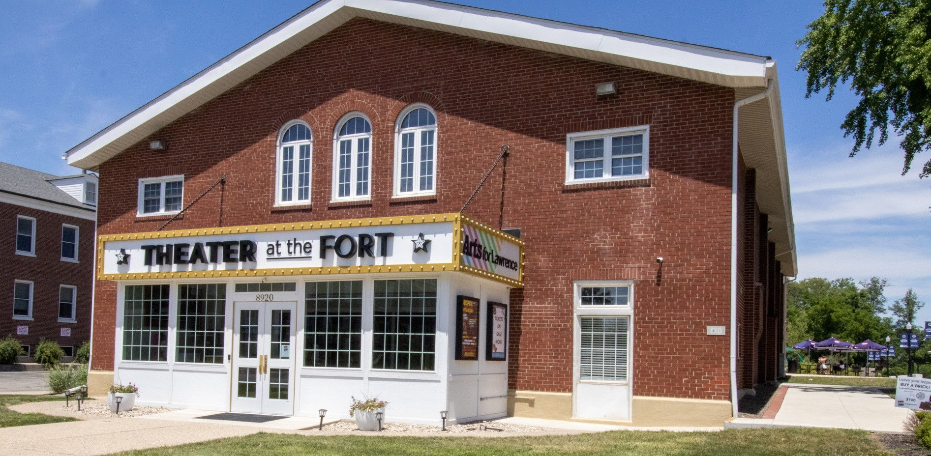 Red brick theater building with large windows, sign "THEATER at the FORT," sunny day, inviting atmosphere, lush lawn.