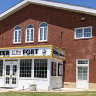 Red brick theater building with large windows, sign "THEATER at the FORT," sunny day, inviting atmosphere, lush lawn.