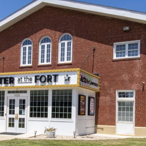Red brick theater building with large windows, sign "THEATER at the FORT," sunny day, inviting atmosphere, lush lawn.