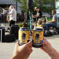 Two people toast with beer cans in a festive outdoor scene, celebrating amid a live band and joyful atmosphere.