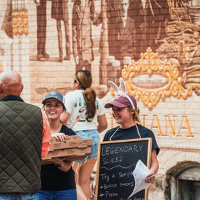 Cheerful people enjoy pizza at a vibrant outdoor market, promoting community and celebration in a lively atmosphere.