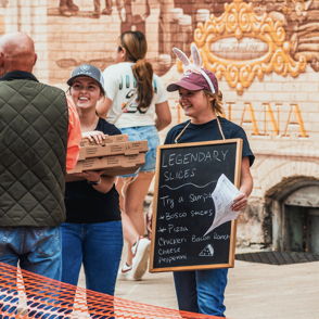 Cheerful people enjoy pizza at a vibrant outdoor market, promoting community and celebration in a lively atmosphere.