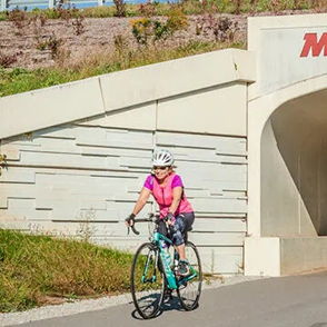 A cyclist in pink rides along a bike path to a "MONON" tunnel, while a dog walker in turquoise enjoys the vibrant scenery.