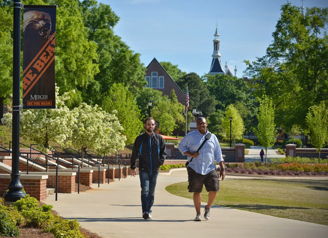 Two individuals walk a sunny path among greenery, enjoying friendship and leisure in a cheerful, tranquil setting.