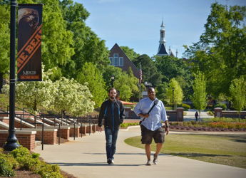 Two individuals walk a sunny path among greenery, enjoying friendship and leisure in a cheerful, tranquil setting.