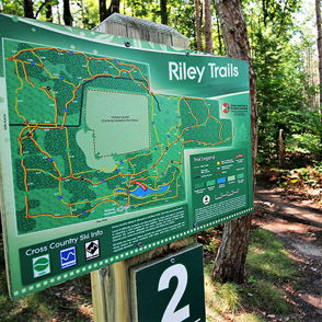 Trail sign for "Riley Trails" in a lush, wooded area, inviting exploration and outdoor activities.