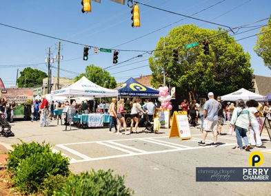 Lively street scene at a community festival with tents, crowds, joy, and inclusivity, all under a bright, sunny sky.