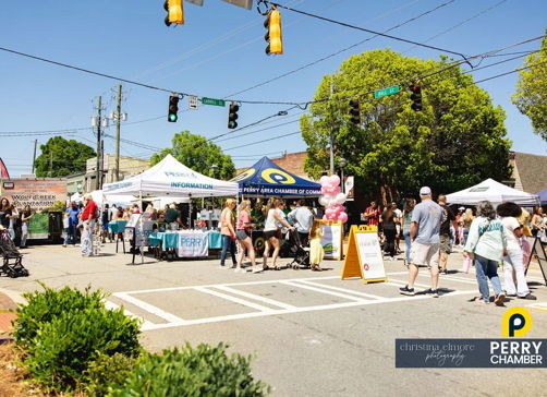 Lively street scene at a community festival with tents, crowds, joy, and inclusivity, all under a bright, sunny sky.