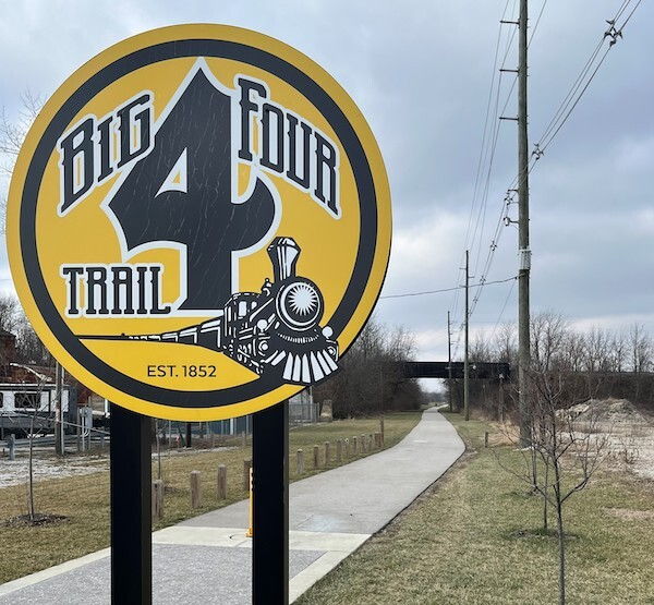 Sign for "Big Four Trail" features a steam locomotive illustration, set against a paved pathway and overcast sky, evoking adventure.