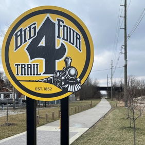 Sign for "Big Four Trail" features a steam locomotive illustration, set against a paved pathway and overcast sky, evoking adventure.